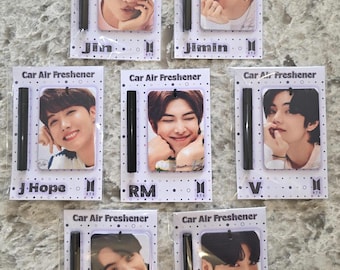 May include: Seven white car air fresheners with black sticks. Each air freshener features a different member of the K-pop group BTS. The names of the members are printed below their photos: Jin, Jimin, J-Hope, RM, V, Suga, and Jung Kook.