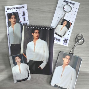 May include: A selection of merchandise featuring a person in a white shirt. Items include a bookmark, a small notepad, a keyring, and a photo card. The keyring and bookmark have the word "Keyring" and "Bookmark" printed on them.