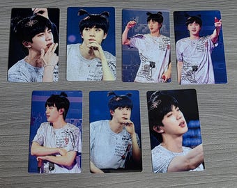 May include: Seven rectangular photo cards featuring a person with dark hair styled in two small buns, wearing a white t-shirt with black graphic text and illustrations. The person is captured in various poses, including holding a microphone and gesturing.
