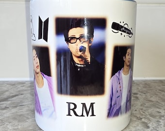 May include: White ceramic mug with a blue rim featuring a collage of photos of a man with the text "RM" printed below the photos. The mug also has a black and white logo of the letter "B" with a cloud and a microphone with the text "Namjoon" printed on the mug.