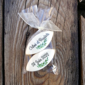 Peut inclure: Deux biscuits blancs en forme d'amande avec des motifs de branches d'olivier vertes et du texte noir. Le biscuit du haut dit "Chloe et Baptiste" et le biscuit du bas dit "28 Juin 2022". Les biscuits sont emballés dans un sac en plastique transparent avec un ruban blanc.