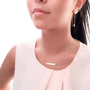 May include: A gold necklace with a thin chain and a small rectangular pendant. The necklace is worn by a person with long dark hair.
