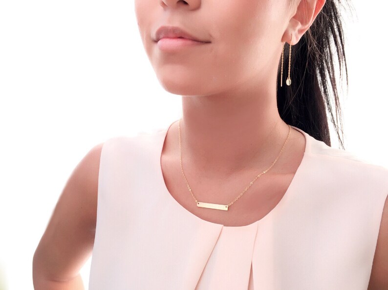 May include: A gold necklace with a thin chain and a small rectangular pendant. The necklace is worn by a person with long dark hair.