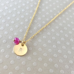 May include: A gold chain necklace with a round gold pendant engraved with the letter "R". A small pink gemstone bead hangs above the pendant.