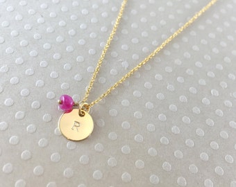 May include: A gold chain necklace with a round gold pendant engraved with the letter "R". A small pink gemstone bead hangs above the pendant.