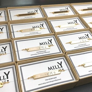 May include: A collection of gold bar bracelets with personalized initials, packaged in small boxes with the brand name "MILLY" printed on them. Each box has a website address printed on it.