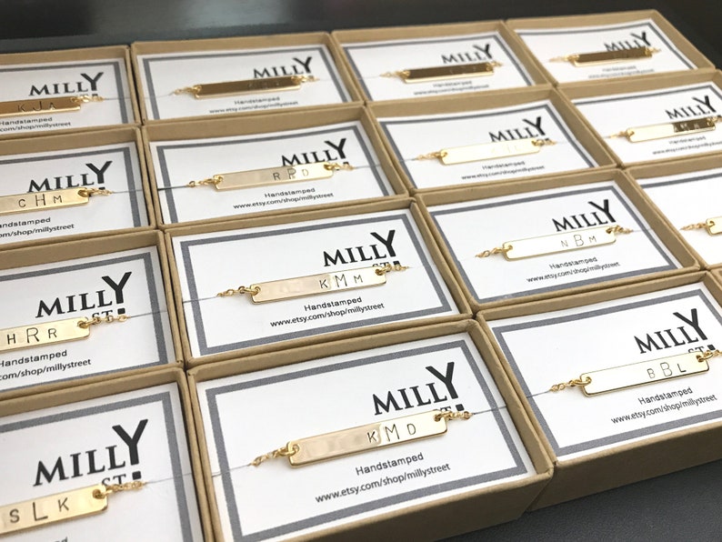 May include: A collection of gold bar bracelets with personalized initials, packaged in small boxes with the brand name "MILLY" printed on them. Each box has a website address printed on it.