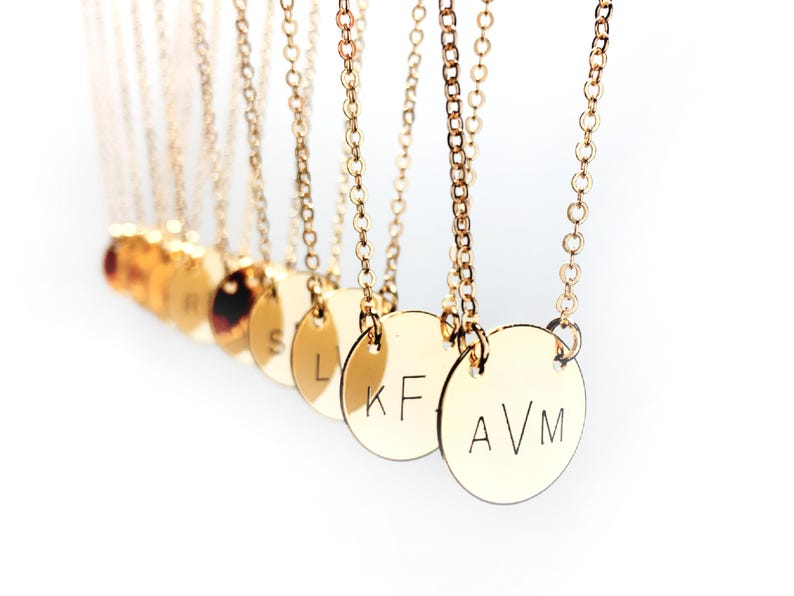 May include: A close-up of several gold necklaces with round pendants. Each pendant has a different letter engraved on it, including "AVM", "KF", "L", and "S".