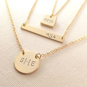 May include: Three gold necklaces with personalized nameplate charms. The top necklace has a square charm with the initials "GMS". The middle necklace has a rectangular bar charm with the initials "MSA". The bottom necklace has a round charm with the initials "SHE".