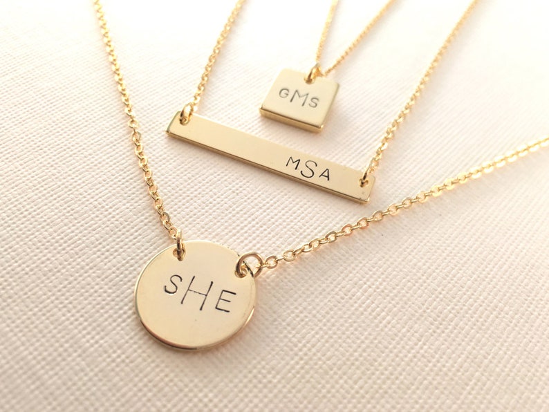 May include: Three gold necklaces with personalized nameplate charms. The top necklace has a square charm with the initials "GMS". The middle necklace has a rectangular bar charm with the initials "MSA". The bottom necklace has a round charm with the initials "SHE".