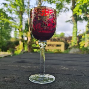 May include: A red wine glass with gold detailing and a decorative pattern. The glass has a clear stem and a round base.