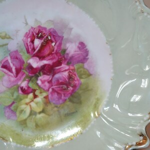 Könnte beinhalten: Ein antiker Porzellanteller mit einem zarten Blumendesign mit rosa Rosen und grünen Blättern. Der Teller hat einen gewellten Rand mit goldener Verzierung.