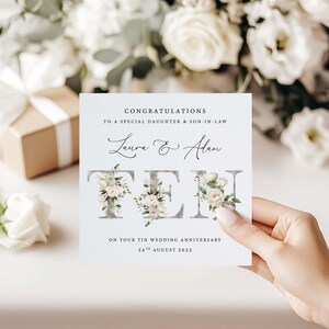 May include: A white card with the text "Congratulations to a special daughter & son-in-law Laura & Adam Ten on your tin wedding anniversary 24th August 2025". The letters of the word "Ten" are decorated with white flowers and green leaves.