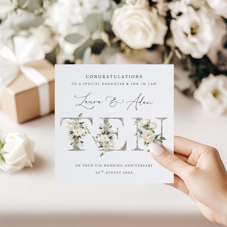 May include: A white card with the text "Congratulations to a special daughter & son-in-law Laura & Adam Ten on your tin wedding anniversary 24th August 2025". The letters of the word "Ten" are decorated with white flowers and green leaves.