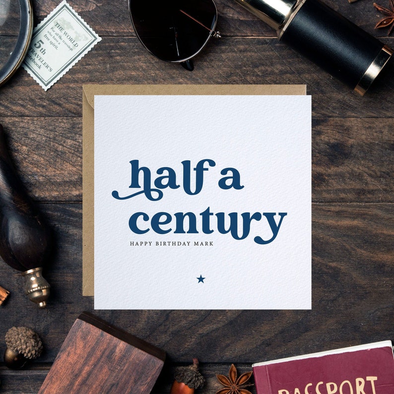 以下が含まれることがあります： 「half a century」と「Happy Birthday Mark」という青い文字が書かれた白いカード。カードは茶色の木製の表面に置かれ、下部に星が描かれている。