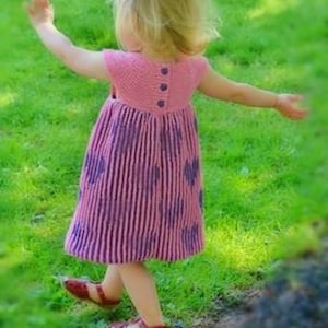 May include: A young child wearing a handmade pink and purple knitted dress with a striped pattern and heart accents. The dress has a button-up back and is paired with red sandals. The child is standing on green grass.