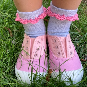 May include: A pair of pink canvas shoes with white soles and laces. The shoes are worn by a person wearing grey socks with pink crocheted trim.