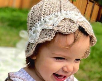 May include: A beige crochet bonnet featuring a textured, speckled yarn construction with a decorative white lace-like trim along the brim. The hat has a rounded crown and a scalloped edge, showcasing intricate hand-stitched details throughout the design.