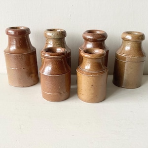May include: A collection of six cylindrical brown stoneware jars with narrow necks and thick, rounded rims. The jars vary slightly in height and shade, ranging from light tan to deep chocolate brown, each featuring a smooth, glazed finish.