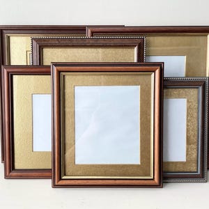 May include: A collection of five rectangular picture frames featuring dark brown wooden borders with gold-toned inner accents and textured gold matting. Each frame contains a central white rectangular insert, arranged in a layered, overlapping display.