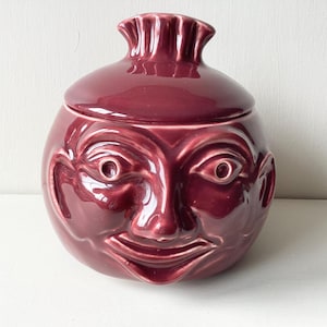 May include: A burgundy ceramic jar shaped like a face with a whimsical expression. The jar has a lid with a decorative top.