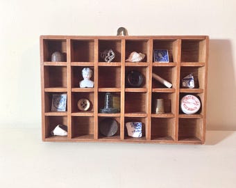 May include: A wooden shadow box with 24 compartments, each holding a different small antique item. Items include a thimble, a seashell, a button, a stamp, and a small porcelain doll. The box has a brass hook for hanging.