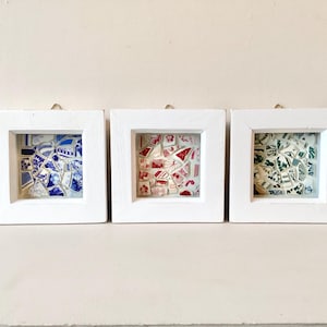 May include: Three square white framed mosaic art pieces. Each frame contains a different colour scheme of broken ceramic pieces. One frame has blue and white, another red and white, and the last green and white.