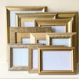 May include: A collection of rectangular picture frames in various shades of gold, featuring smooth, textured, and ornate embossed border designs. The frames are stacked together, each containing a blank white rectangular insert for display purposes.