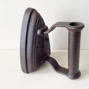 May include: A dark brown, cast iron flat iron with a handle. The iron has a rounded base and a handle that curves upwards. The handle features a circular opening at the top. This iron is likely an antique or a decorative item.