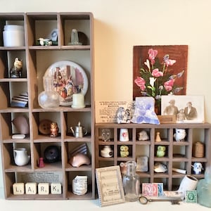 May include: Two wooden display shelves filled with vintage items, such as figurines, a small teapot, a plate, and a floral painting. The word "LARK" is spelled out with letter tiles.