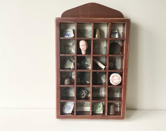 May include: A vintage wooden shadow box with a dark brown finish, divided into multiple compartments. Each compartment contains a different antique item, including a doll head, thimbles, and small ceramic pieces. The background features a faded black and white image.