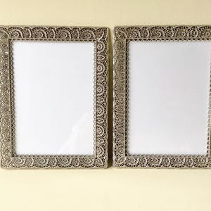 May include: Two rectangular silver picture frames with ornate, lacy edges. The frames have a vintage aesthetic and feature a white interior space for photos or artwork. The frames are displayed side by side.