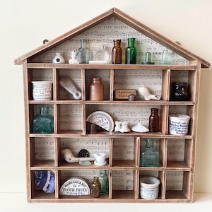May include: A house-shaped wooden shelf unit displaying a collection of antique bottles, jars, and curios. The back of the shelf is lined with sheet music. The unit is constructed from light brown wood.