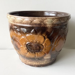 May include: A brown ceramic flower pot with a floral design. The pot has a wide, round shape with a textured surface and a dark brown rim. The floral pattern is in shades of brown and tan.