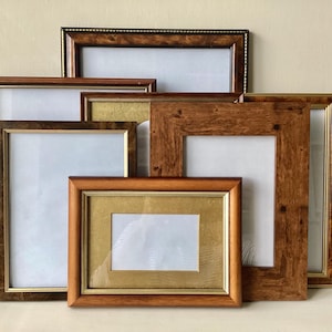 May include: A collection of six rectangular picture frames arranged in a layered stack. The frames feature various shades of brown wood grain finishes, some with gold-toned inner trim accents, surrounding white matting and empty centre spaces.