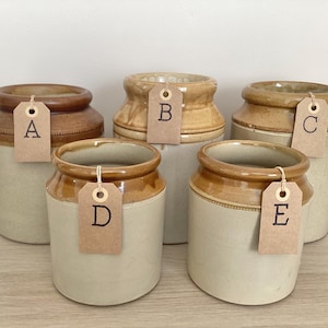 May include: Five cylindrical ceramic stoneware jars with a two-tone design featuring a light cream-coloured base and a darker brown glazed rim. Each jar has a brown paper tag attached with a string, labelled with the letters A, B, C, D, and E.