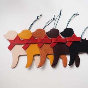 May include: Five dog-shaped ornaments in a row, each with a red scarf and green string for hanging. The dogs are in shades of beige, yellow, orange, brown, and black. These ornaments are likely for a Christmas tree or other holiday decor.