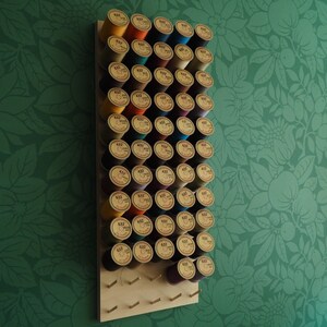 May include: A wooden wall-mounted storage rack with 48 spools of thread in various colors, including orange, red, blue, purple, and yellow. The spools are arranged in rows on the rack.
