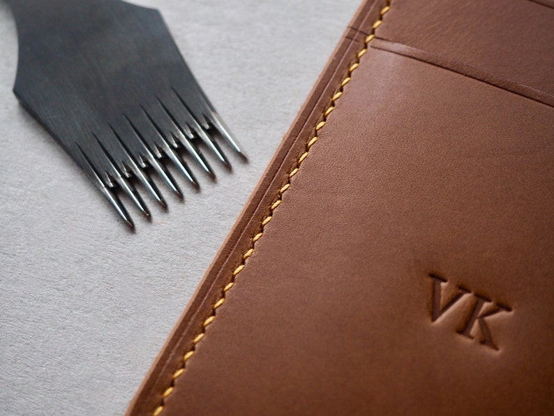 May include: Close-up of a brown leather wallet with yellow stitching and the initials "VK" embossed on the front. A leather stitching tool is visible in the background.