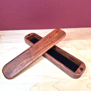 May include: A long, rectangular wooden box with a rich reddish-brown grain pattern. The box consists of a base lined with black velvet and a matching lid that rests diagonally across it, featuring small circular magnetic closures at each end.