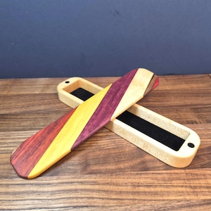 May include: A rectangular wooden box with a sliding lid featuring diagonal stripes of purple, yellow, and light tan wood. The interior of the box is lined with black felt, and the lid is positioned diagonally across the open base.
