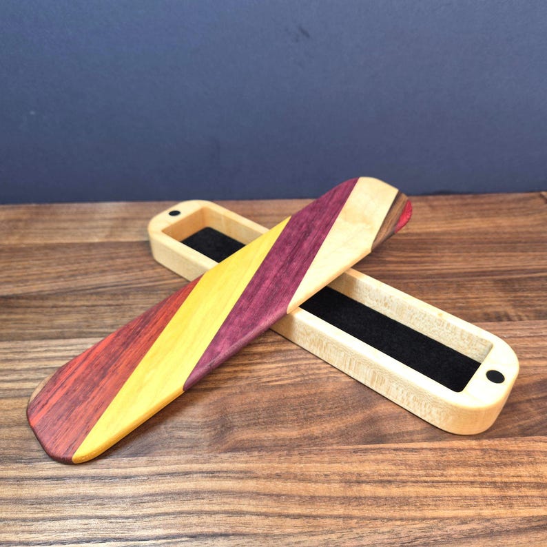 May include: A rectangular wooden box with a sliding lid featuring diagonal stripes of purple, yellow, and light tan wood. The interior of the box is lined with black felt, and the lid is positioned diagonally across the open base.