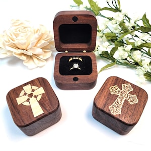 May include: Three wooden ring boxes, one open with a diamond ring inside. The boxes are square with rounded corners and feature gold-coloured cross designs. The open box has a black velvet interior.