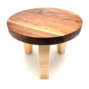 May include: A small, round wooden plant stand with a dark brown and light brown top and three light-colored legs. The stand is made of natural wood and has a simple, minimalist design. Perfect for displaying small plants or decorative items.