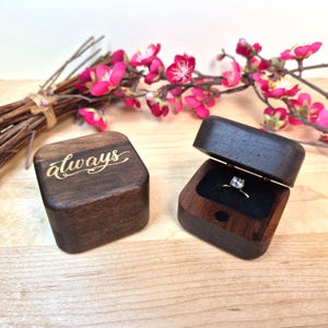 May include: Two square dark wood ring boxes with rounded corners. One box is closed and features the word always engraved in gold script on the lid. The other box is open, revealing a silver ring with a clear stone set in a black velvet insert.
