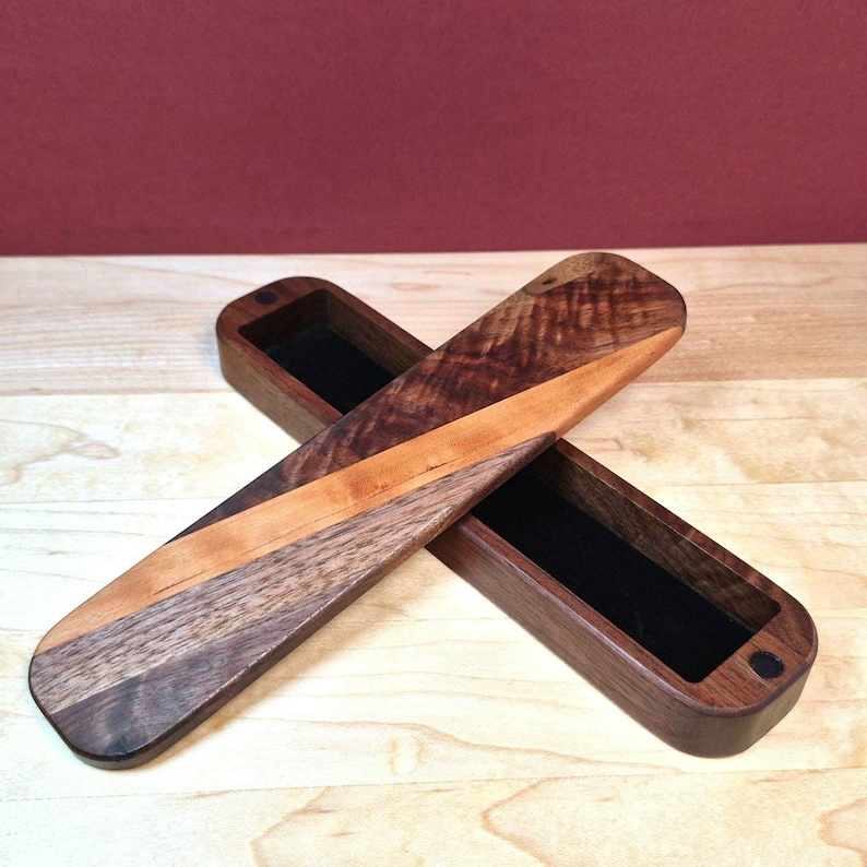 May include: A long, rectangular wooden box with a lid, featuring a dark brown wood grain and a diagonal light brown wood inlay. The interior of the box is lined with black velvet, and the lid is positioned diagonally across the open base.