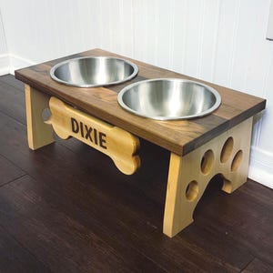 May include: A raised pet feeder with a dark brown wooden top and two stainless steel bowls. The light-colored wooden base features a bone-shaped cutout on the front with the name DIXIE engraved in dark letters and paw print cutouts on the sides.