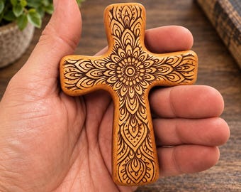 May include: A light brown wooden cross featuring a dark brown laser-engraved mandala pattern. The cross has rounded edges and a smooth finish, displaying intricate symmetrical floral-like details that radiate from the center of the object.