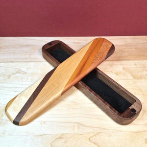 May include: A long, rectangular wooden case with a swivel lid featuring diagonal stripes of light, medium, and dark brown wood. The interior of the case is lined with black felt, and the lid is secured by a small, circular black magnetic closure.