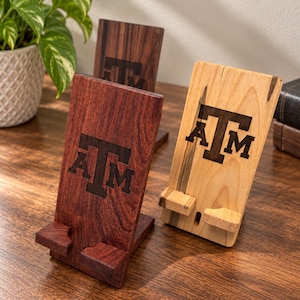 May include: Two wooden phone stands with a rectangular base and a support ledge. One stand is made of dark reddish-brown wood, and the other is light tan wood with dark grain. Both feature the laser-engraved ATM logo on the front surface.